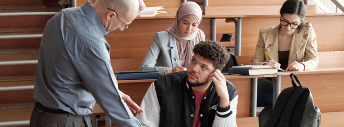 Teacher speaking to student at students' desk in a college classroom.
