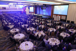 Large banquet hall filled with round tables and people attending a formal event. A stage with speakers and a large screen displaying Annual Meeting is at the front. Plates and glasses indicate a meal is being served.