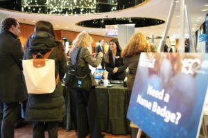 A woman at a registration desk assists attendees at an event. A sign in the foreground reads Need a Name Badge? and features an 150th Anniversary logo. The scene is in a hotel or conference center lobby with warm lighting and a circular ceiling.