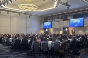 A large group of people seated at round tables in a conference room during an annual meeting, with presentation screens displaying event information.