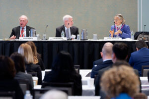 Three panelists sit at a table speaking to an audience during a conference. One woman gestures while talking, and two men listen. Nameplates and microphones are visible on the table. Audience members face the panelists.