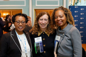 Three women stand side-by-side at a professional event, smiling for the camera. Each wears a name tag on a lanyard. The background shows a room with a branded step-and-repeat backdrop featuring the 150 logo of the New York State Bar Association.