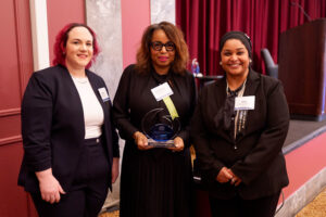 Three women in business attire pose indoors. The woman in the center holds a glass award and wears glasses. All three are smiling and standing in front of a stage with red curtains.