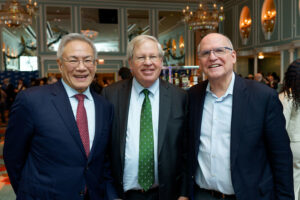 Three men in suits smile and pose together at an indoor event with elegant chandeliers and large mirrors in the background. Other attendees can be seen mingling behind them.
