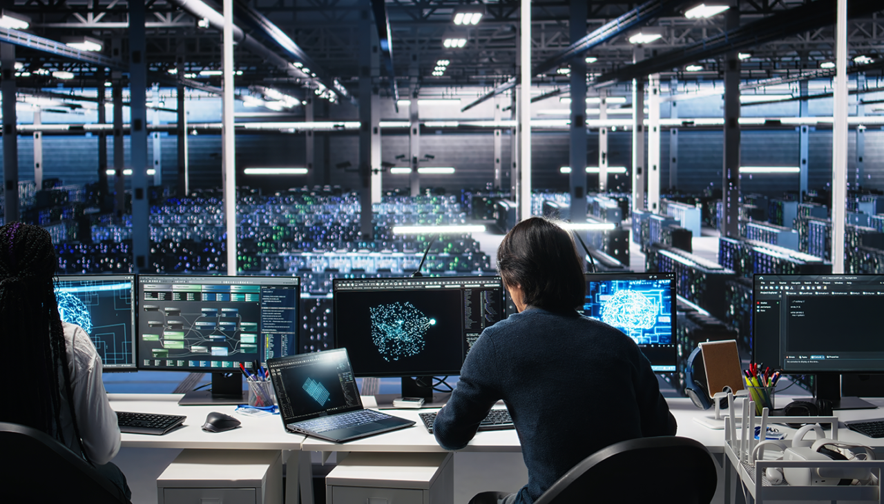 Two people sit at desks with multiple monitors displaying data and code, overlooking a large, modern, high-tech server room filled with illuminated server racks. The environment is bright and organized.