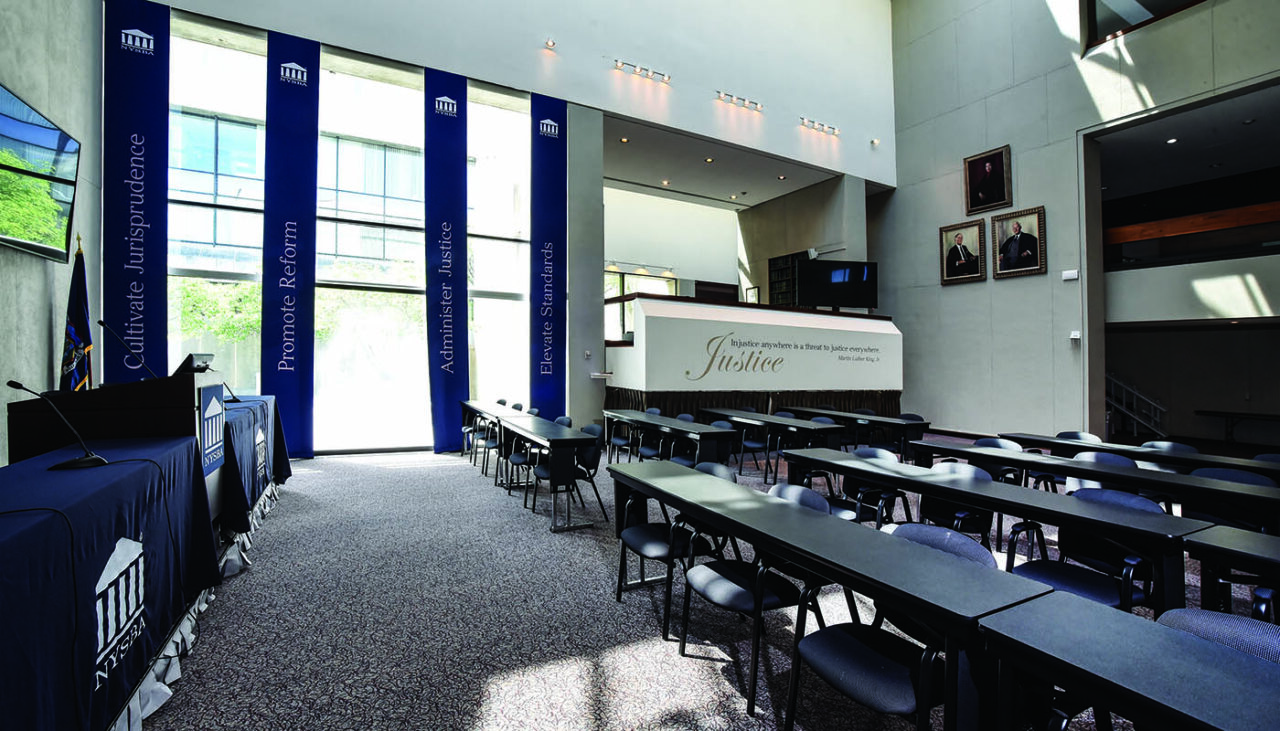 A modern lecture hall with rows of black tables and chairs, high ceilings, large windows, blue banners with text, and framed portraits on the gray wall. Sunlight streams in, creating a bright, open atmosphere.