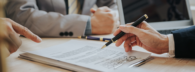 Three people in business attire sit at a table, reviewing and discussing a contract agreement. One person points at the document while another holds a pen, ready to sign. A laptop and pens are also on the desk.