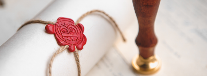 A rolled parchment tied with twine and sealed with red wax lies on a document, next to a wooden-handled stamp.