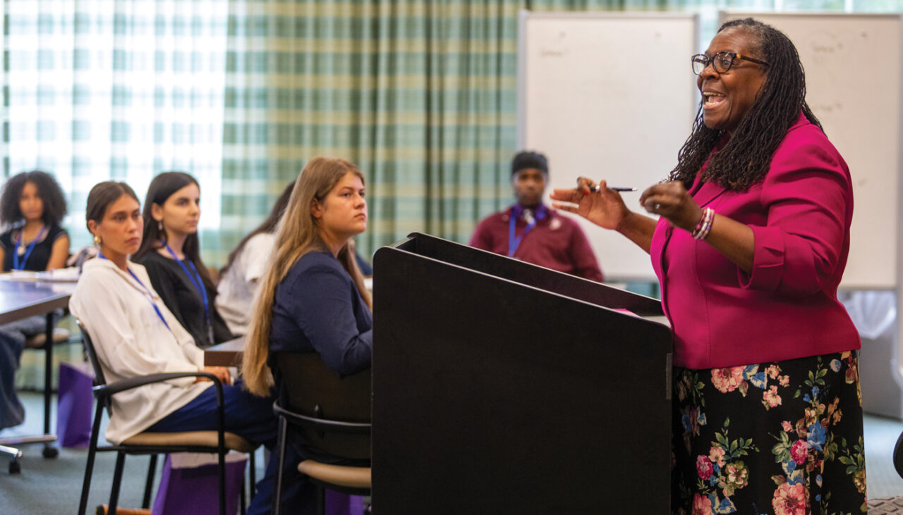 Female instructor standing at a classroom podium addressing a group of teenage students.