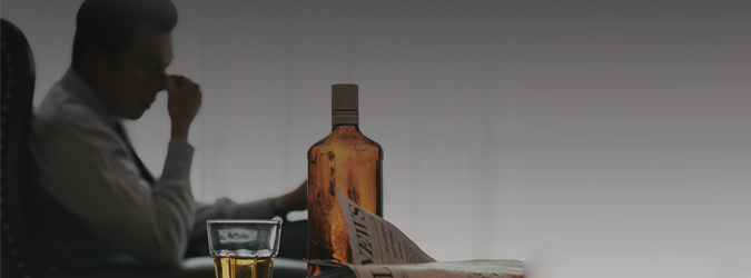 A man sits in the background holding his face, looking stressed. In the foreground, there is a whiskey bottle, a glass of whiskey, and a folded newspaper on a table.
