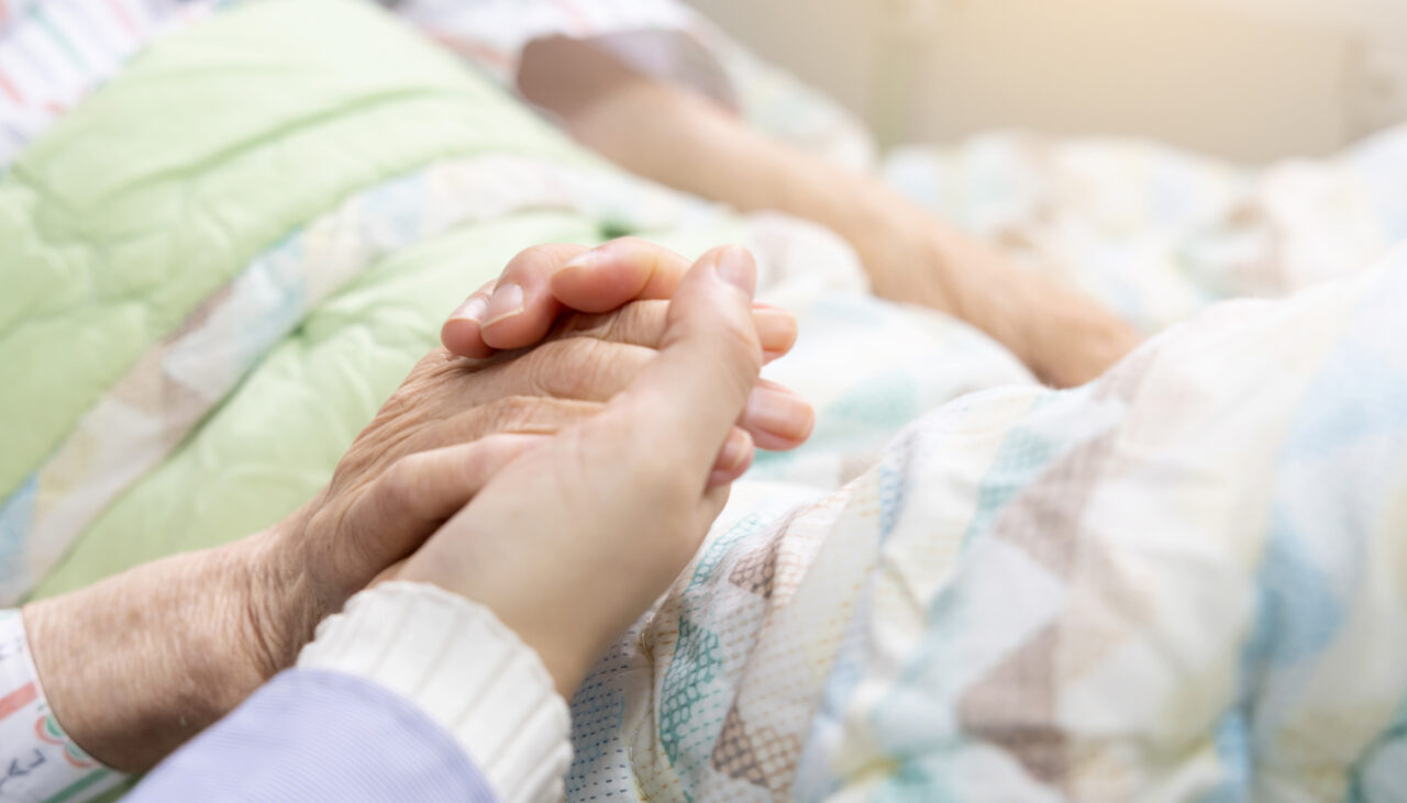 A close-up of a younger person gently holding the hand of an elderly person, who is lying in bed under a pastel-colored quilt, suggesting comfort and support.