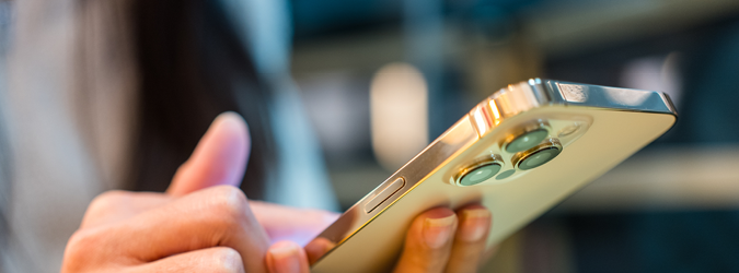 A person holds a smartphone with one hand and uses the other hand to touch the screen. The phones camera lenses are visible, and the background is blurred.