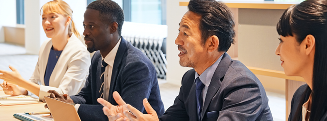 Four professionals in business attire sit at a conference table, engaged in discussion. Two men and two women appear focused and attentive, with papers and a laptop in front of them in a modern office setting.