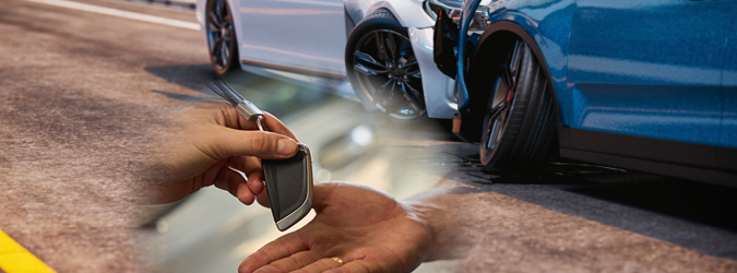 A hand holds a car key fob in the foreground, while two cars with visible front-end damage from a collision are shown in the background on a street.