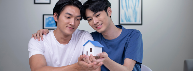 Two people sit close together, smiling and holding a small model house, suggesting a sense of partnership or shared dreams about home ownership. Framed art is visible on the wall behind them.
