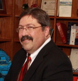 A man with glasses, a mustache, and dark hair is wearing a black suit, white shirt, and red tie. He is smiling and sitting in front of a wooden bookshelf filled with books and awards.