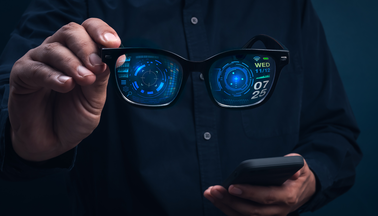 A person holds smart glasses displaying digital interfaces and information, while using a smartphone. The glasses show futuristic graphics, including a clock and date, against a dark background.