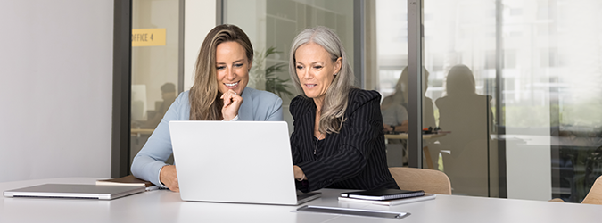 Two women sit at a table, smiling and looking at a laptop screen together. They appear to be collaborating in a modern office setting with notebooks and a closed laptop on the table.