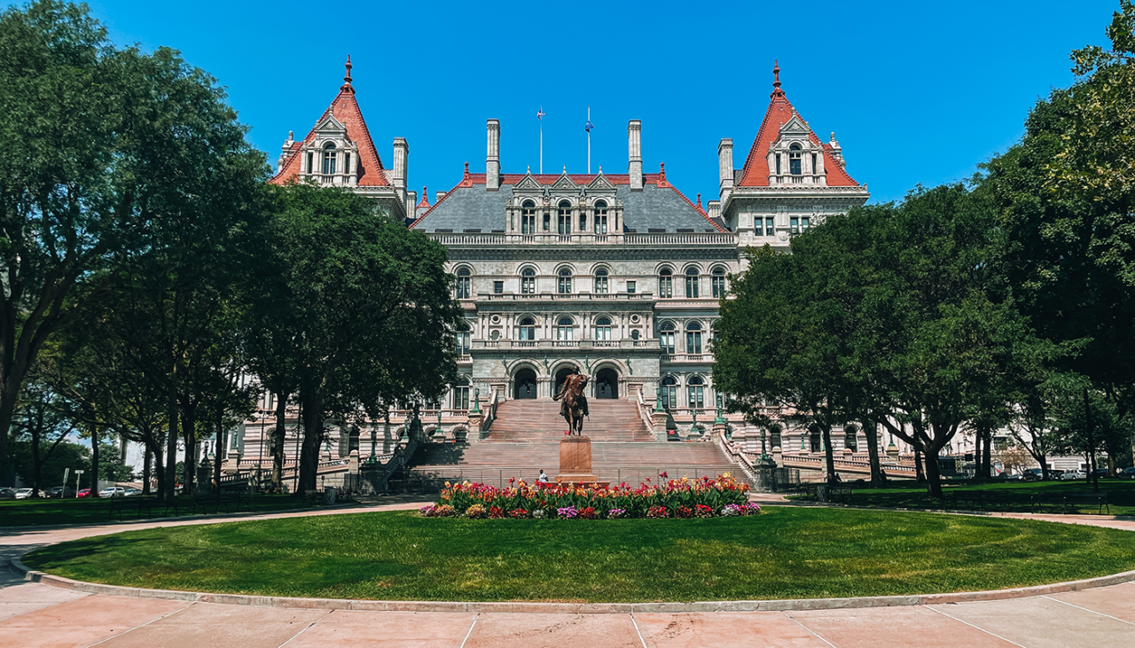 New York State Capitol, A grand stone building with red-roofed towers stands behind a statue and steps, framed by green trees and a circular lawn with flowers, under a clear blue sky.