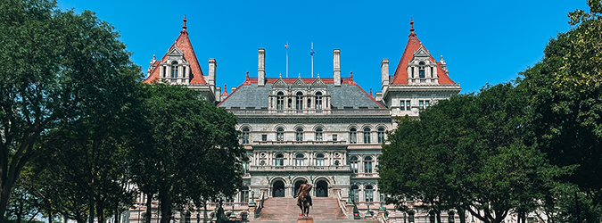 A grand historic building with red-roofed towers and ornate architecture stands behind a staircase, framed by green trees under a clear blue sky.