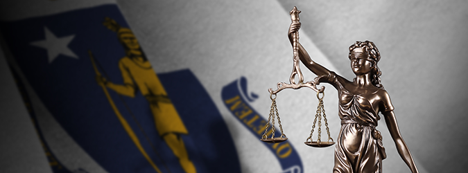 A bronze Lady Justice statue holding scales is in front of a blurred Massachusetts state flag.