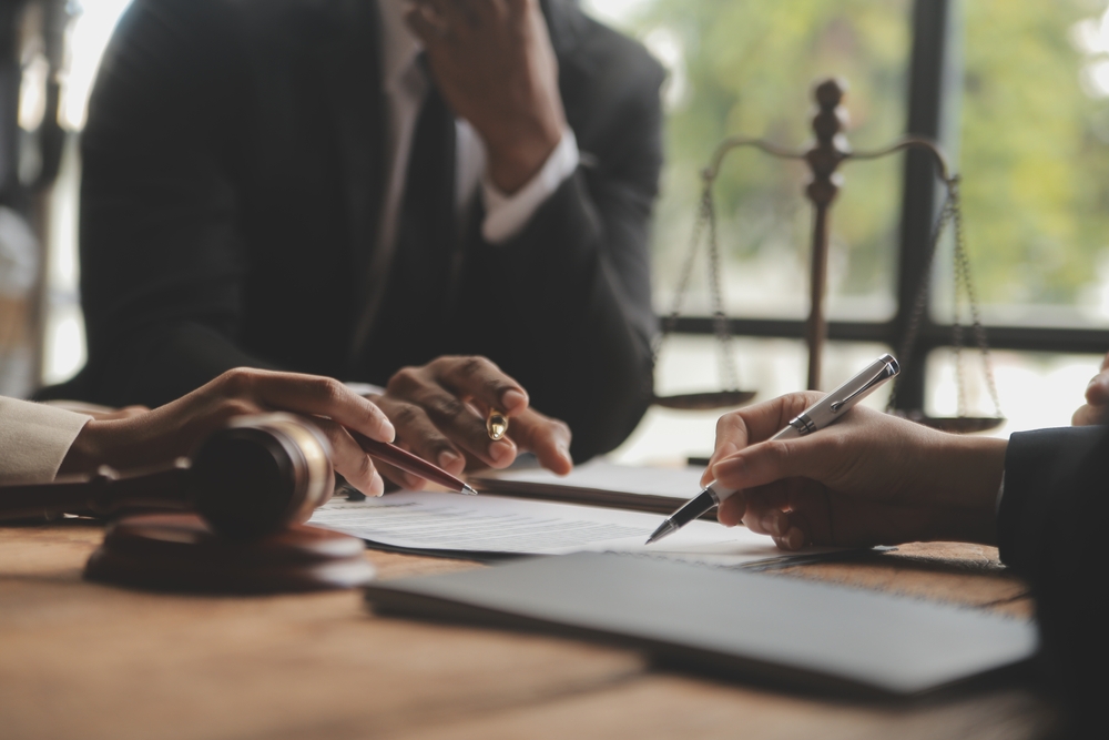 Three people sit at a table with legal documents, a gavel, and scales of justice. One person signs paperwork while others observe and discuss, suggesting a legal or business meeting.