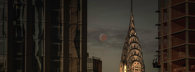 The moon rises behind the illuminated spire of the Chrysler Building, framed by dark glass windows and an unfinished building at dusk.