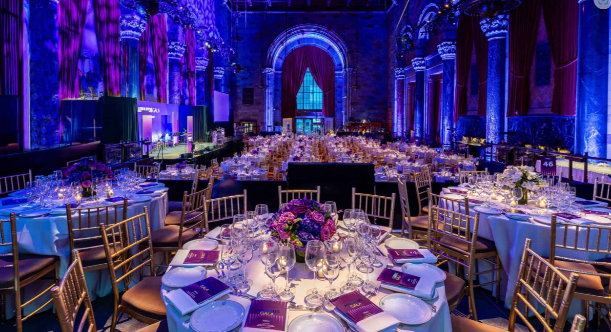 A grand banquet hall with round tables set with flowers, glassware, and purple menus. Gold chairs surround the tables, and the room is lit with purple lighting and large arched windows in the background.