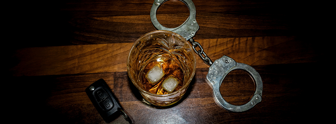A whiskey glass with ice, a car key, and handcuffs are placed on a wooden table, symbolizing the dangers of drinking and driving.