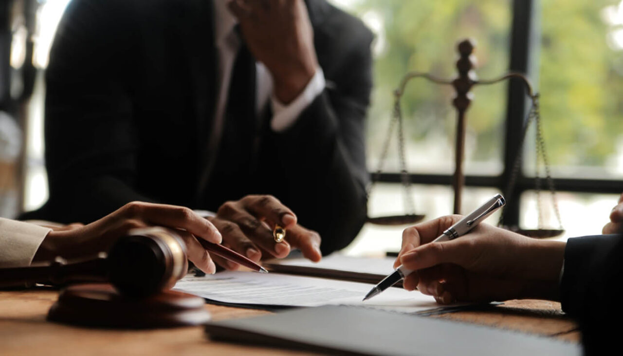 Three people in formal attire discuss and sign legal documents at a desk with a gavel, a pen, and a set of scales visible, suggesting a legal or courtroom setting.