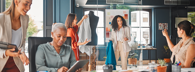 Five women collaborate in a modern office, discussing clothing designs. One presents fabric on a mannequin while others use tablets and phones. Mood boards and fabric samples are on display.