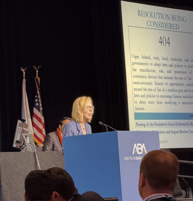 A woman speaks at a podium with an American Bar Association sign. An American flag is behind her, and a projection screen displays a resolution numbered 404. The audience listens attentively.