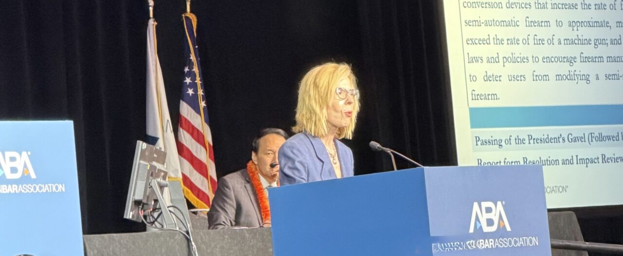 A woman speaks at a podium during an ABA event. Behind her, a projected slide shows Resolution Being Considered 404 about firearm laws. Two flags and another person seated are visible on stage.