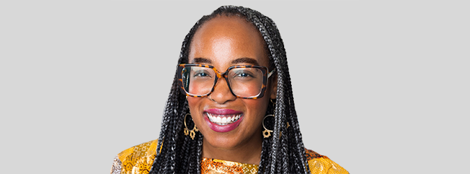 Smiling person with braided hair, wearing large glasses and gold earrings, dressed in a yellow patterned top, in front of a plain light gray background.