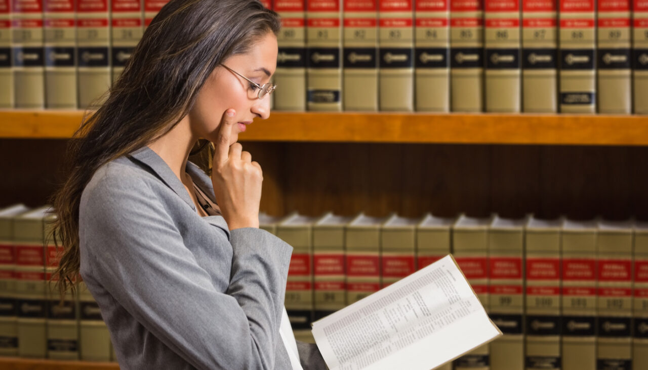 A woman in a gray suit reads a book thoughtfully in front of shelves filled with legal books.