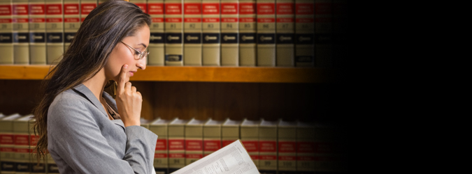 A woman in a gray suit and glasses reads a book, standing in front of shelves filled with legal books.