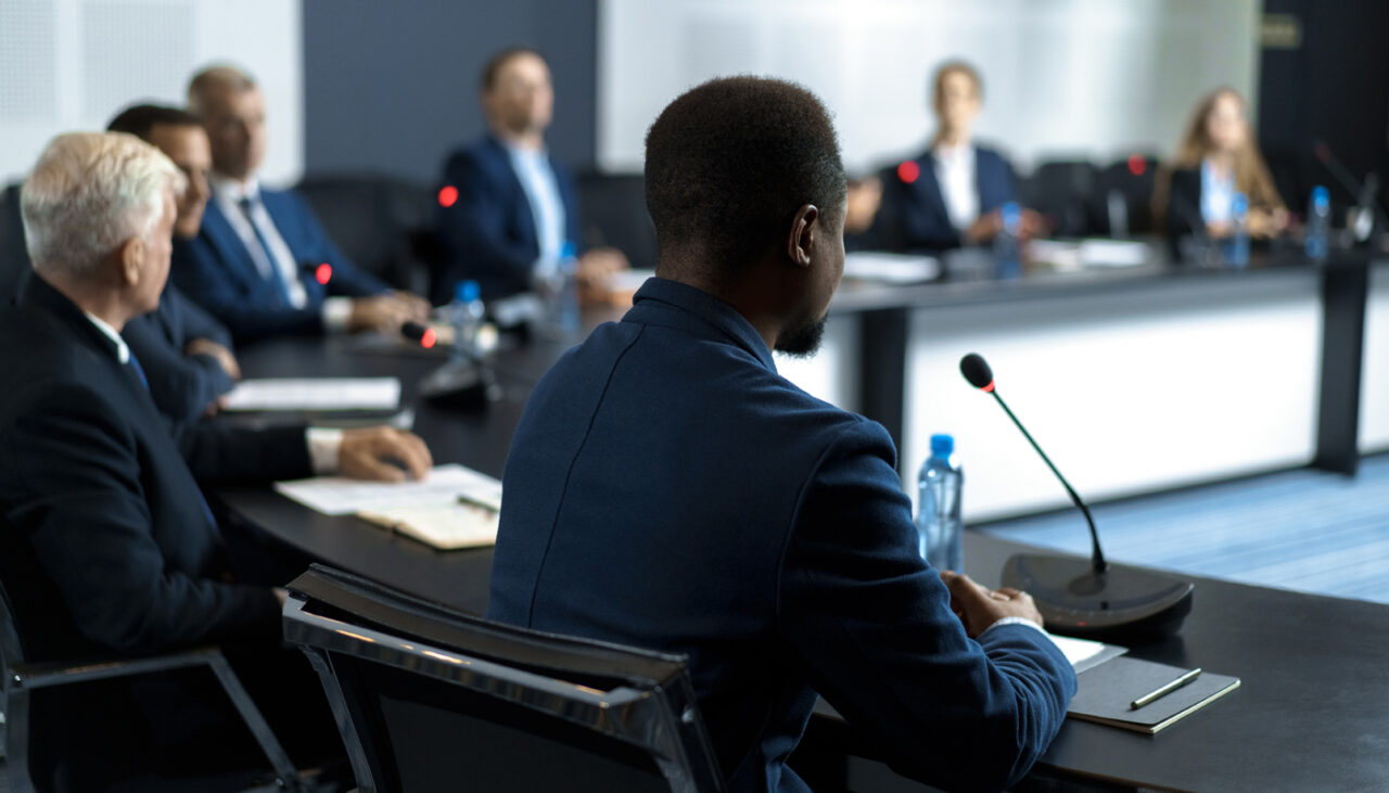 People in business attire sit around a conference table with microphones, participating in a formal meeting or discussion. The focus is on a man in the foreground.