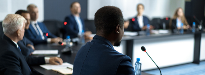 A group of people in business attire sit around a conference table with microphones, engaged in a formal meeting or discussion.