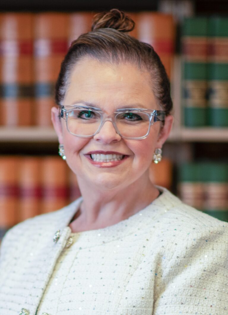 Woman with glasses and earrings, wearing a white textured jacket, smiles in front of a background of bookshelves filled with books. Her hair is neatly pulled back into a bun.
