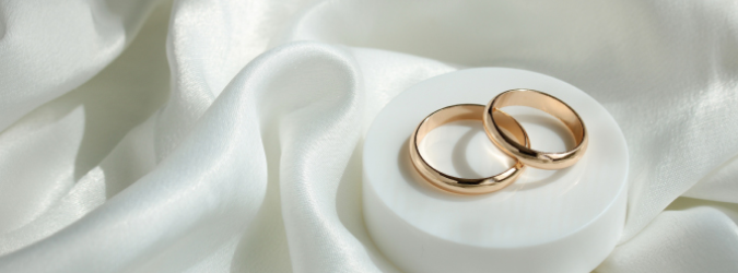 Two gold wedding rings rest on a white round platform, placed on smooth, white satin fabric with gentle folds.