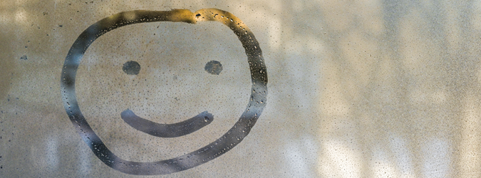 A smiley face is drawn with a finger on a foggy glass window, with blurred light and shadows in the background.