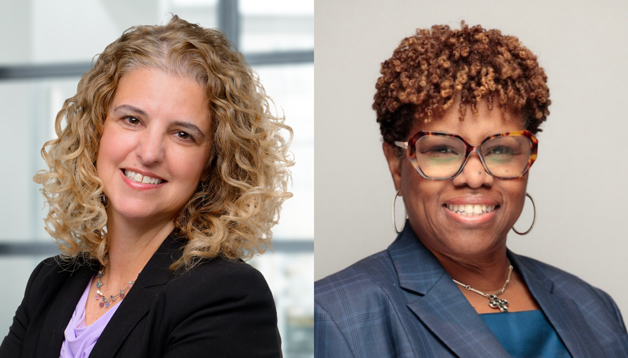 Two women smiling side by side; the left has curly blonde hair, light skin, wearing a black blazer; the right has short curly hair, dark skin, glasses, and a blue jacket.