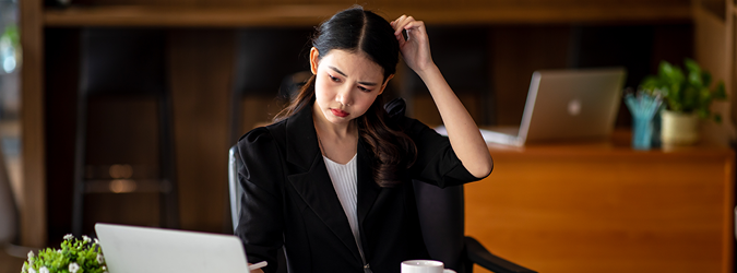 A woman in business attire sits at a desk with a laptop, looking at the screen with a confused expression while scratching her head. An open notebook, coffee cup, and another laptop are visible in the background.