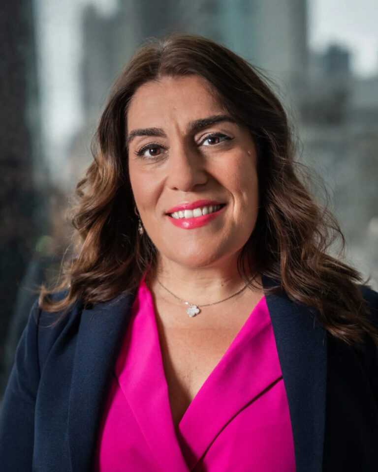 A woman with wavy brown hair, wearing a dark blazer over a bright pink top, smiles at the camera. She has a delicate necklace and is posed indoors with a blurred cityscape background.