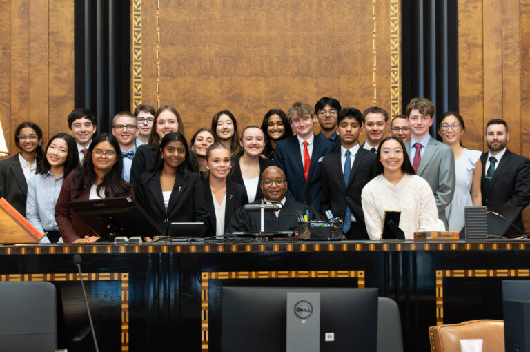 A group of students in formal attire poses together in a courtroom, with one student seated at the judges desk and others standing behind, all smiling at the camera.