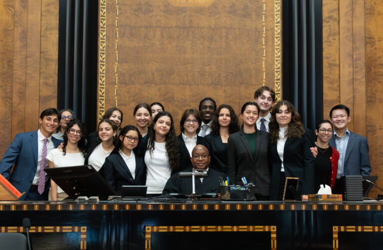 A group of people in formal attire stand behind a judge seated at a large, ornate desk in a courtroom, smiling for a group photo.