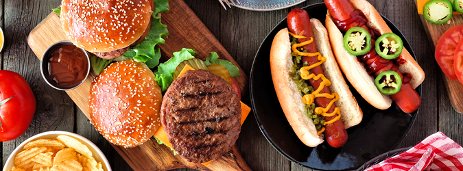 Two cheeseburgers with lettuce and tomato on a wooden board, two hot dogs with toppings on a black plate, potato chips, tomatoes, and sliced pickles on a rustic table.