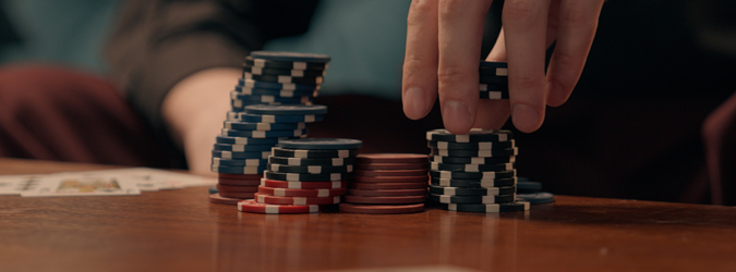 A persons hand is stacking poker chips on a wooden table, with several piles of chips in different colors. Playing cards are partially visible in the corner of the image.