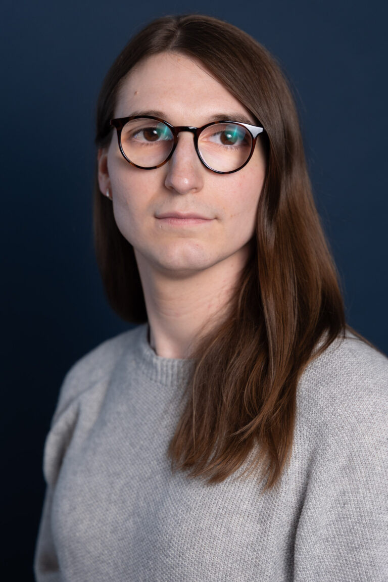 A person with long brown hair and round glasses, wearing a gray sweater, looks slightly to the left against a dark blue background.