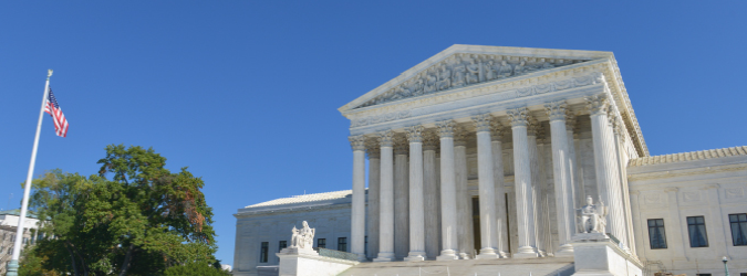 The United States Supreme Court building with tall columns under a clear blue sky and an American flag on a pole in the foreground.